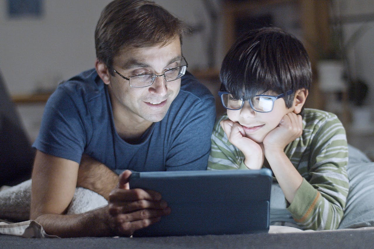 Home A father and son lying in bed, happily watching a tablet indoors at night.